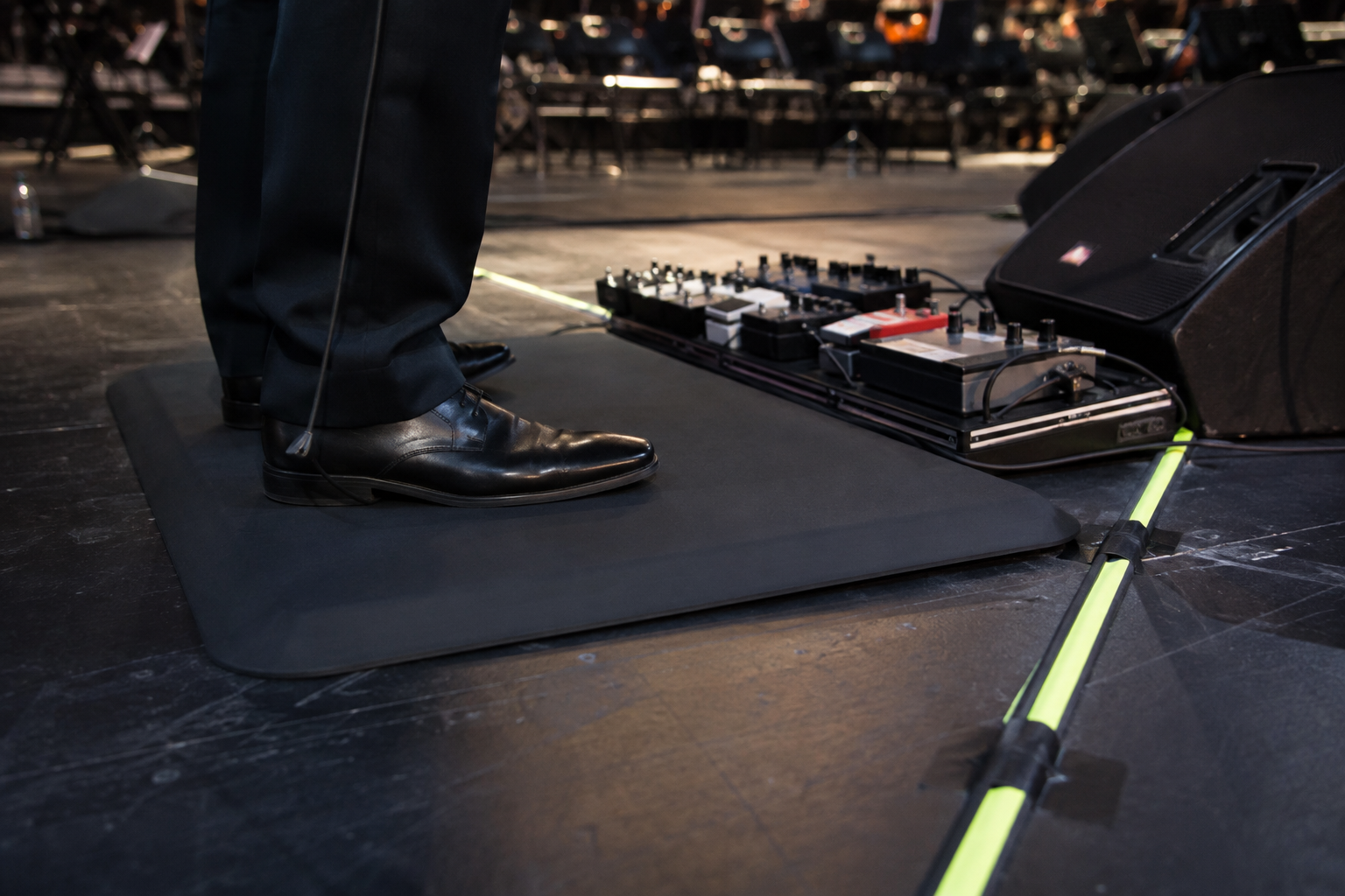 Formal shoes on a beveled anti-fatigue mat with electric violin pedalboard and taped cable route safely beyond the edge