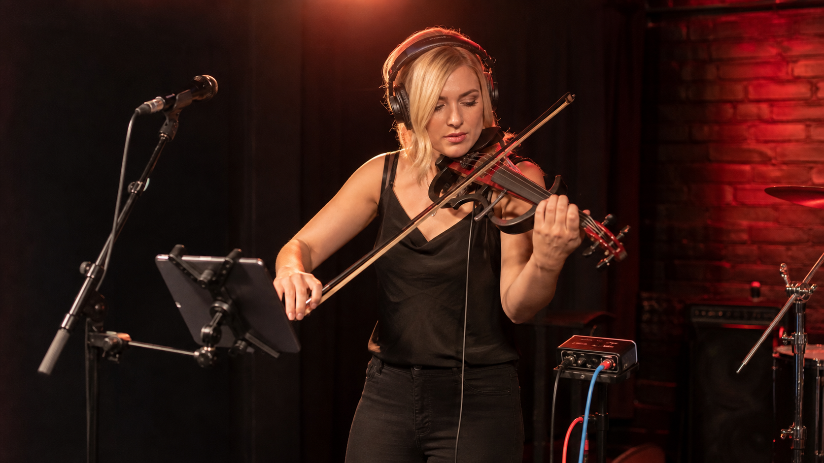 Electric violinist on a small venue stage wearing closed-back headphones during a rehearsal with tablet mount and DI box