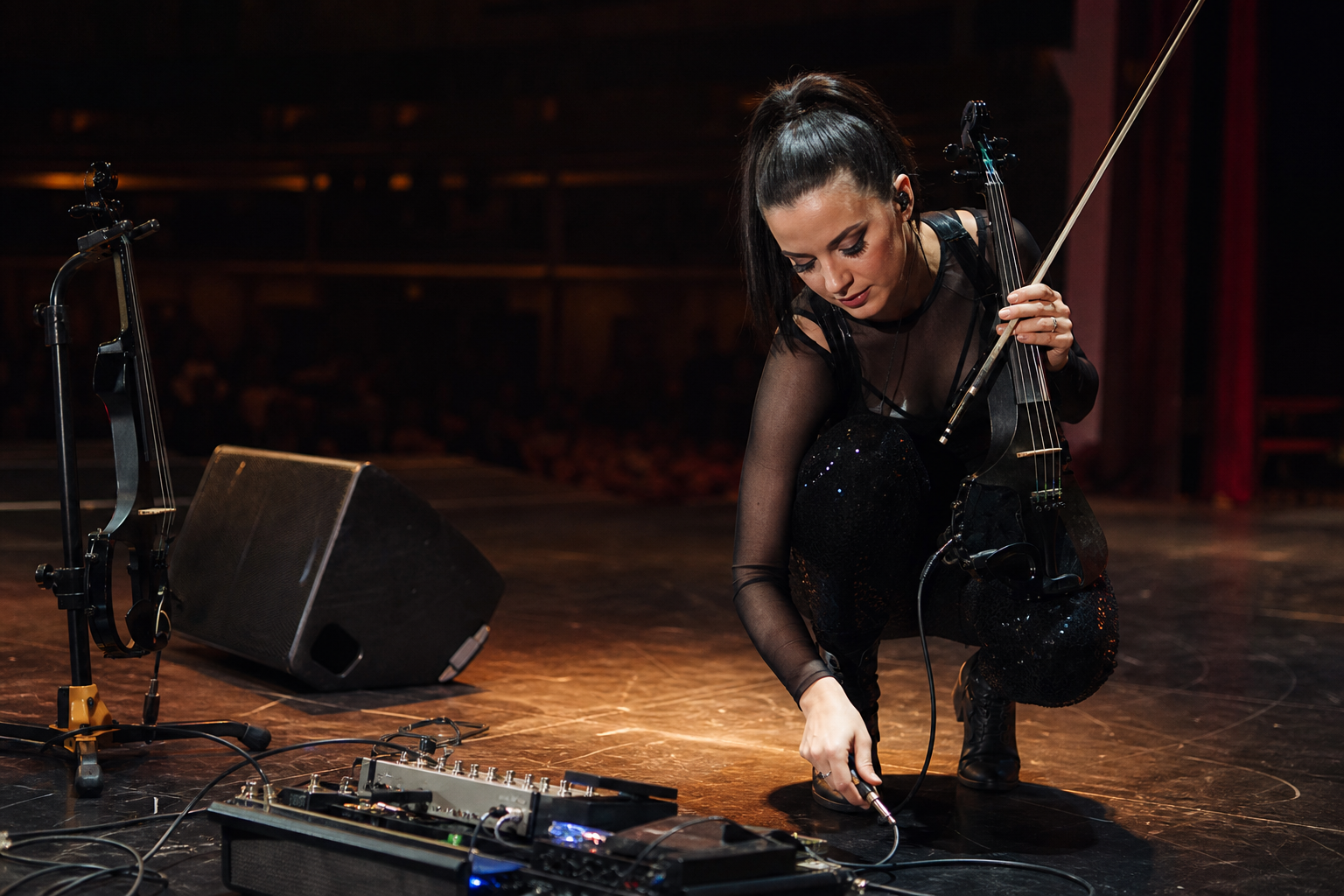 Electric violin performer reconnecting an instrument cable near a pedalboard during a quiet stage reset
