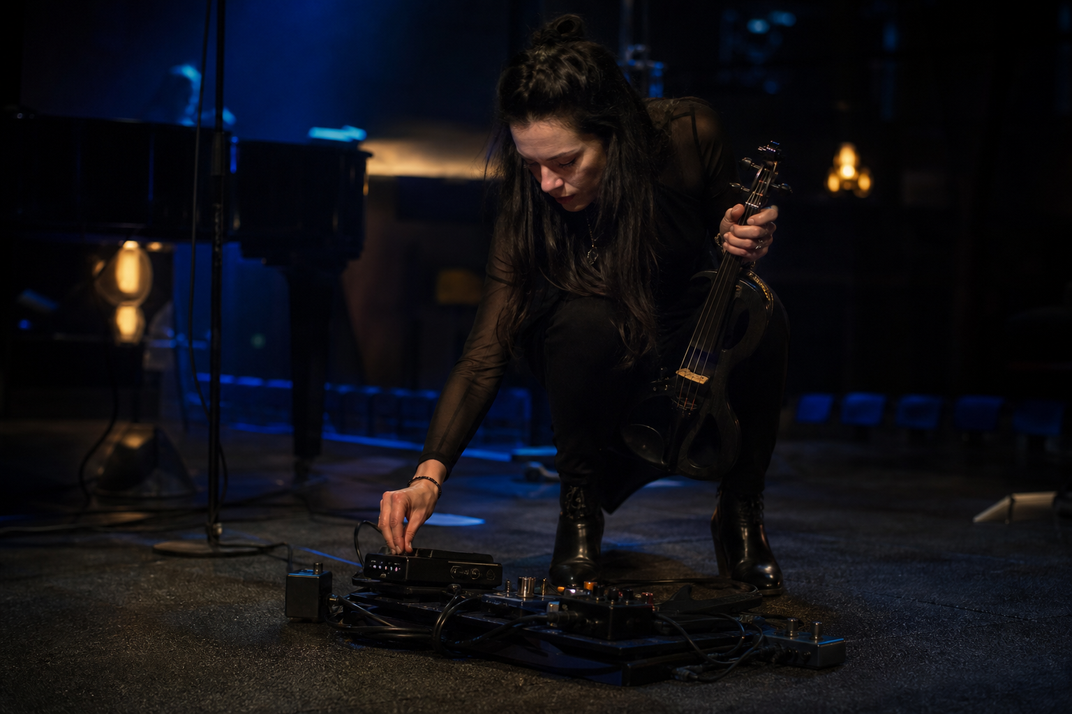 Изпълнителка на електрическа цигулка е клекнала до тих pedalboard и проверява power индикаторите след soundcheck в тъмна зала